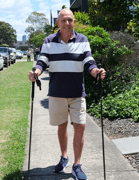 image 7 Clover Canes Older man walking outdoors with two trekking poles on a sidewalk wearing a striped polo shirt and khaki shorts smiling