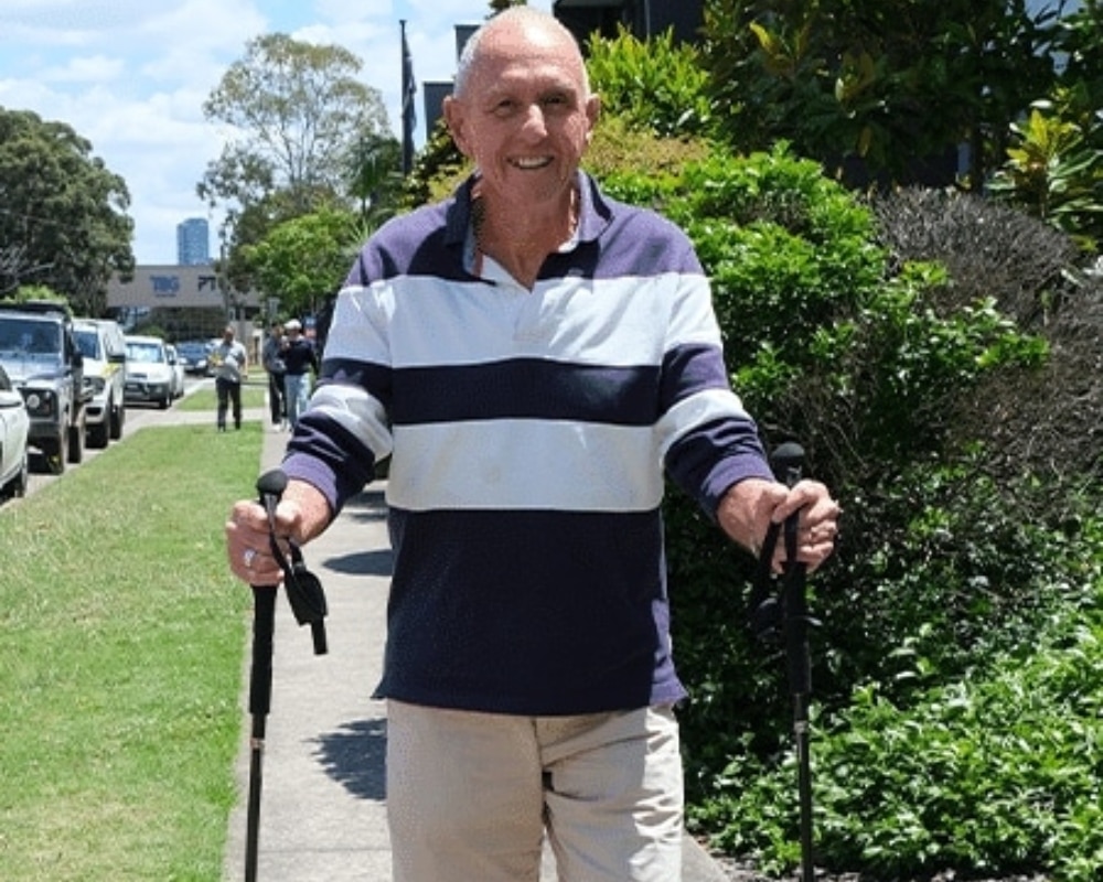 Older man walking on a sidewalk with trekking poles, smiling, wearing a navy and white striped sweater and khaki shorts.
