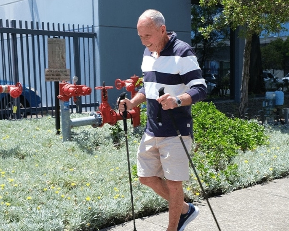 Older man walking with trekking poles beside a red fire hydrant and fence on a sunny day in a landscaped area near a building.